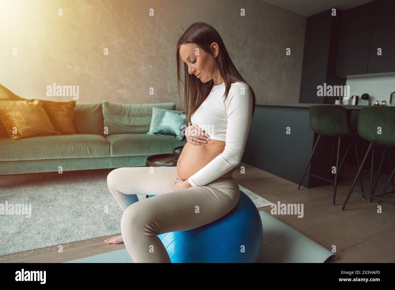 Calmare la donna incinta seduta su una palla da fitness e toccando la pancia. Madre felice che fa esercizi fisici di supporto per una gravidanza liscia. Foto Stock