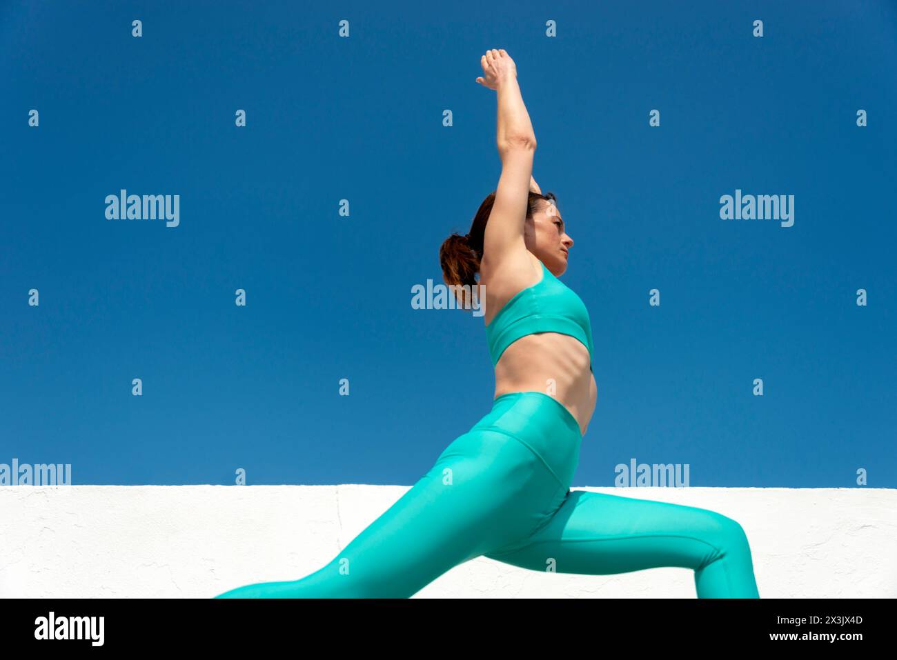 Vista laterale di una donna che indossa abbigliamento sportivo verde che fa esercizio di yoga con sfondo blu del cielo, posa di affondo alto yoga o Anjaneyasana, Foto Stock