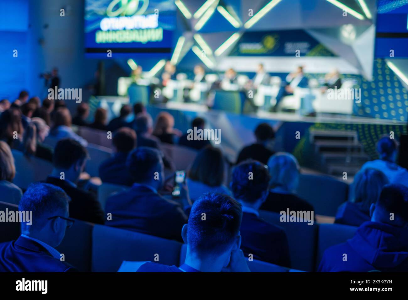 I membri del pubblico in abbigliamento formale si concentrano in una vivace conferenza d'affari, coinvolgendo le discussioni in un ambiente moderno illuminato da una luce blu dinamica. Foto Stock