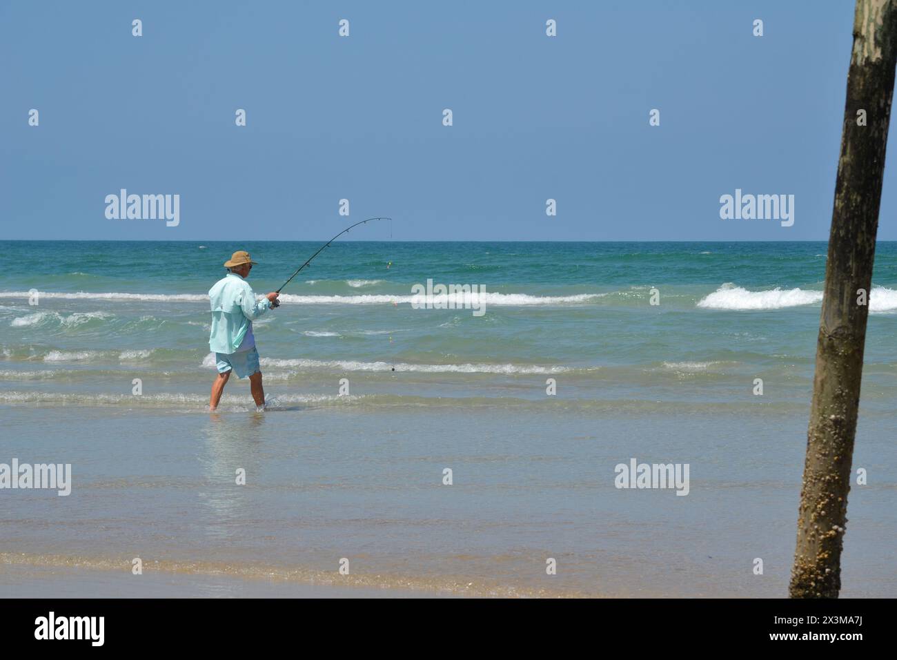 Un pescatore da surf, con un cappello da abbronzatura e una camicia in acqua, entra nell'oceano tenendo la canna da pesca, a Ponce Inlet, Jetty Beach in Florida. Foto Stock