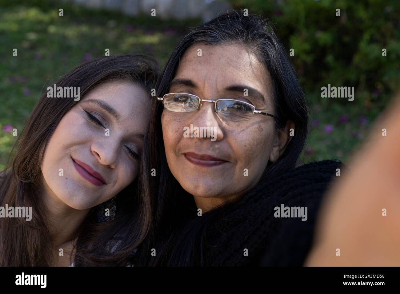 Madre latinoamericana (50) e figlia (24) fanno un selfie nel parco, la figlia si appoggia amorevolmente sulla spalla di sua madre chiudendo gli occhi. Madre" Foto Stock