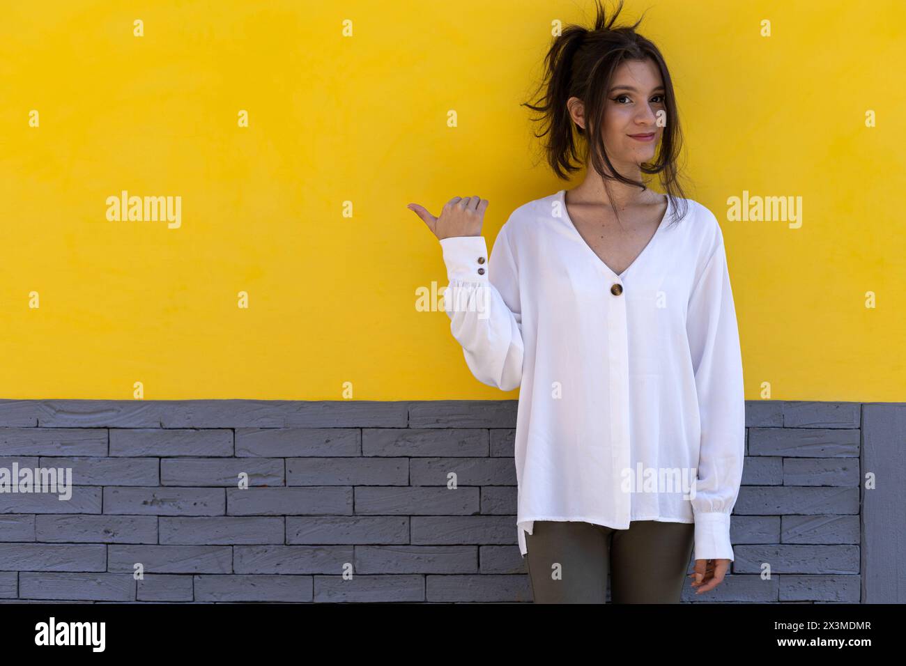 Ritratto di una giovane donna latinoamericana sorridente (24) isolata su sfondo giallo e grigio che punta con il pollice verso uno spazio vuoto. Promozione concettuale Foto Stock