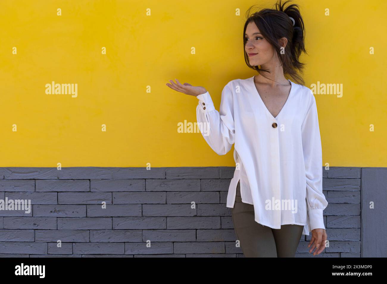Ritratto di una giovane donna latinoamericana sorridente (24) isolata su sfondo giallo e grigio che punta con il palmo della mano a uno spazio vuoto. Promo concettuale Foto Stock