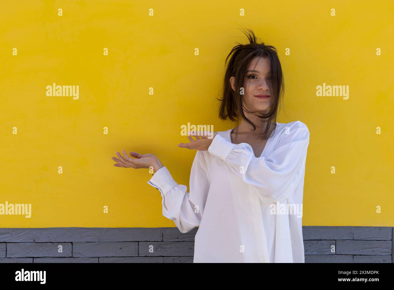 Ritratto di una giovane donna latinoamericana sorridente (24) isolata su sfondo giallo e grigio con palmo di una mano e dito dell'altra mano Foto Stock