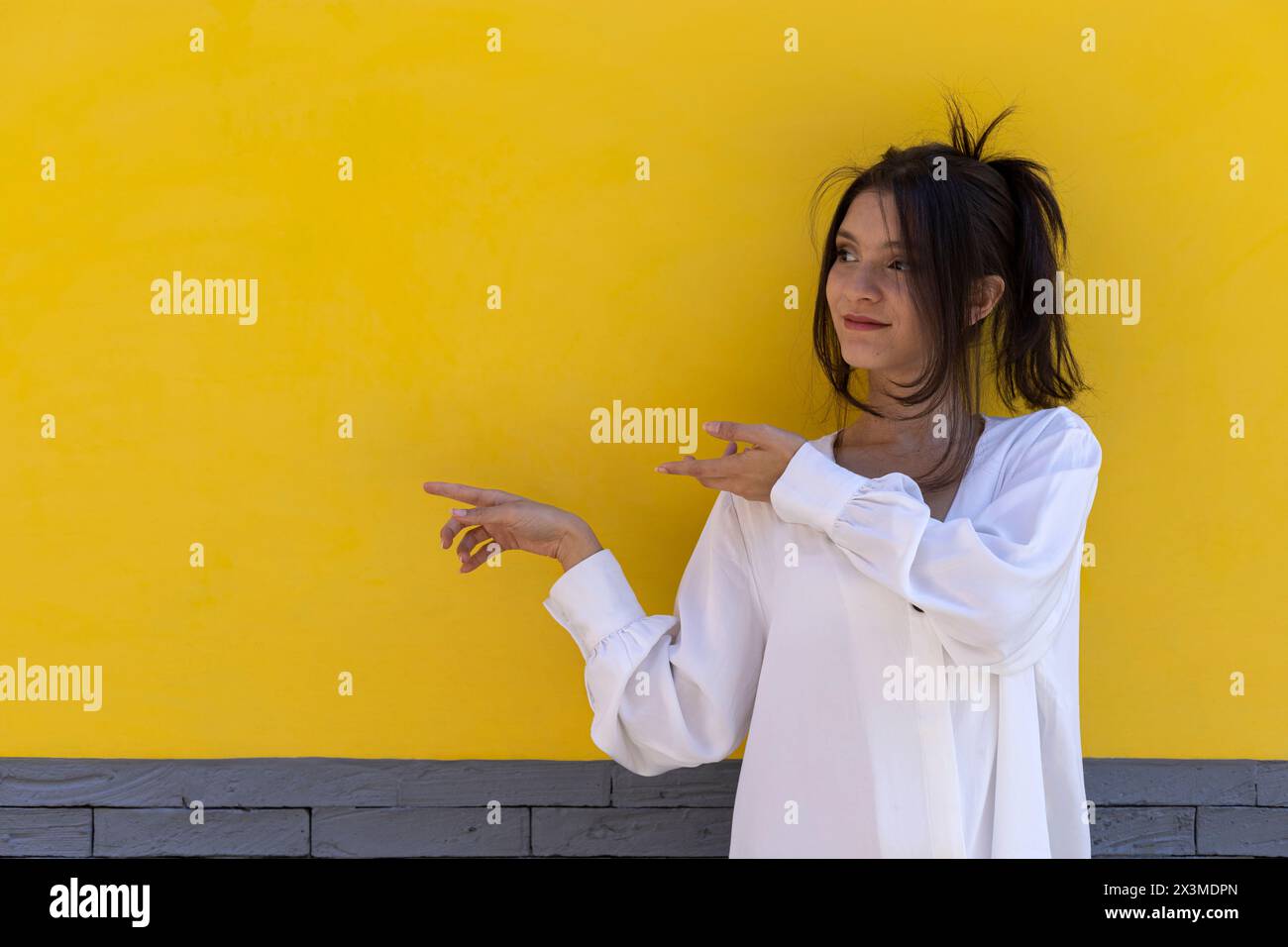 Ritratto di una giovane donna latinoamericana sorridente (24) isolata su sfondo giallo e grigio punta con entrambe le dita della mano verso uno spazio vuoto. Concep Foto Stock