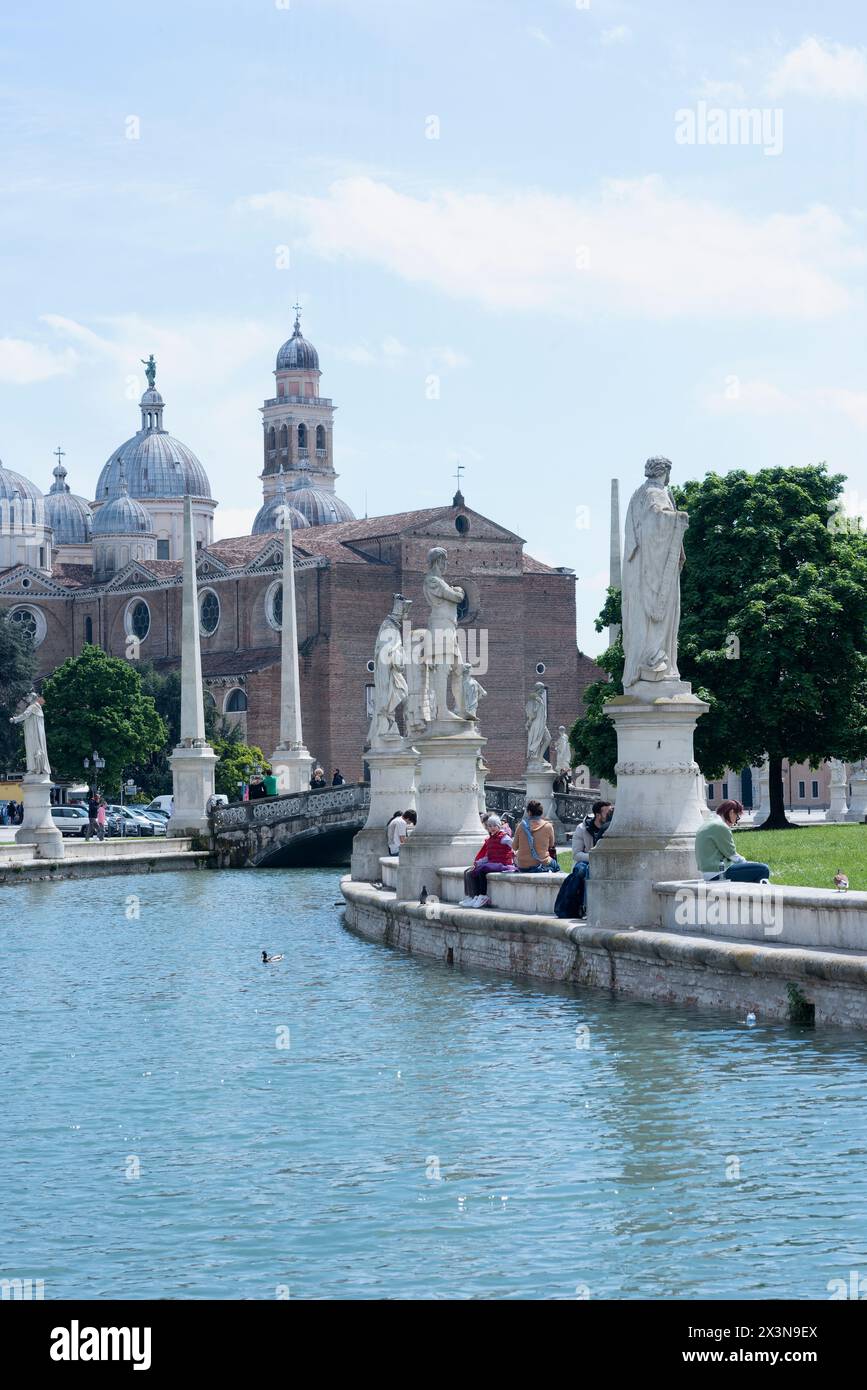Giornata di sole a Prato della Valle con gente sulla riva del canale. L'isola verde centrale, Memia, circondata da statue. In vista della basilica abbaziale di sa Foto Stock