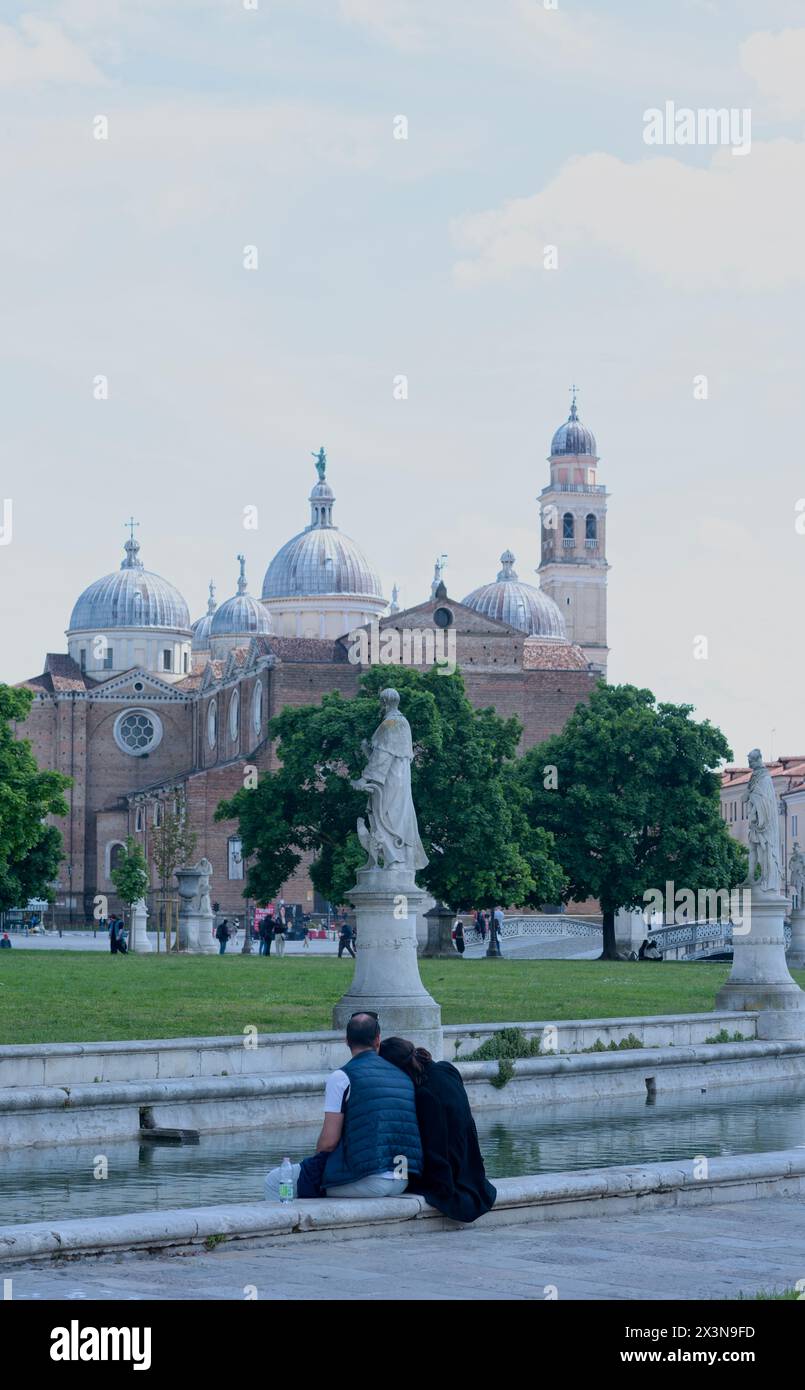 Due amanti seduti sulla riva del canale in una giornata di sole a Prato della Valle, l'isola verde centrale, Memia, circondata da statue. Sullo sfondo Foto Stock