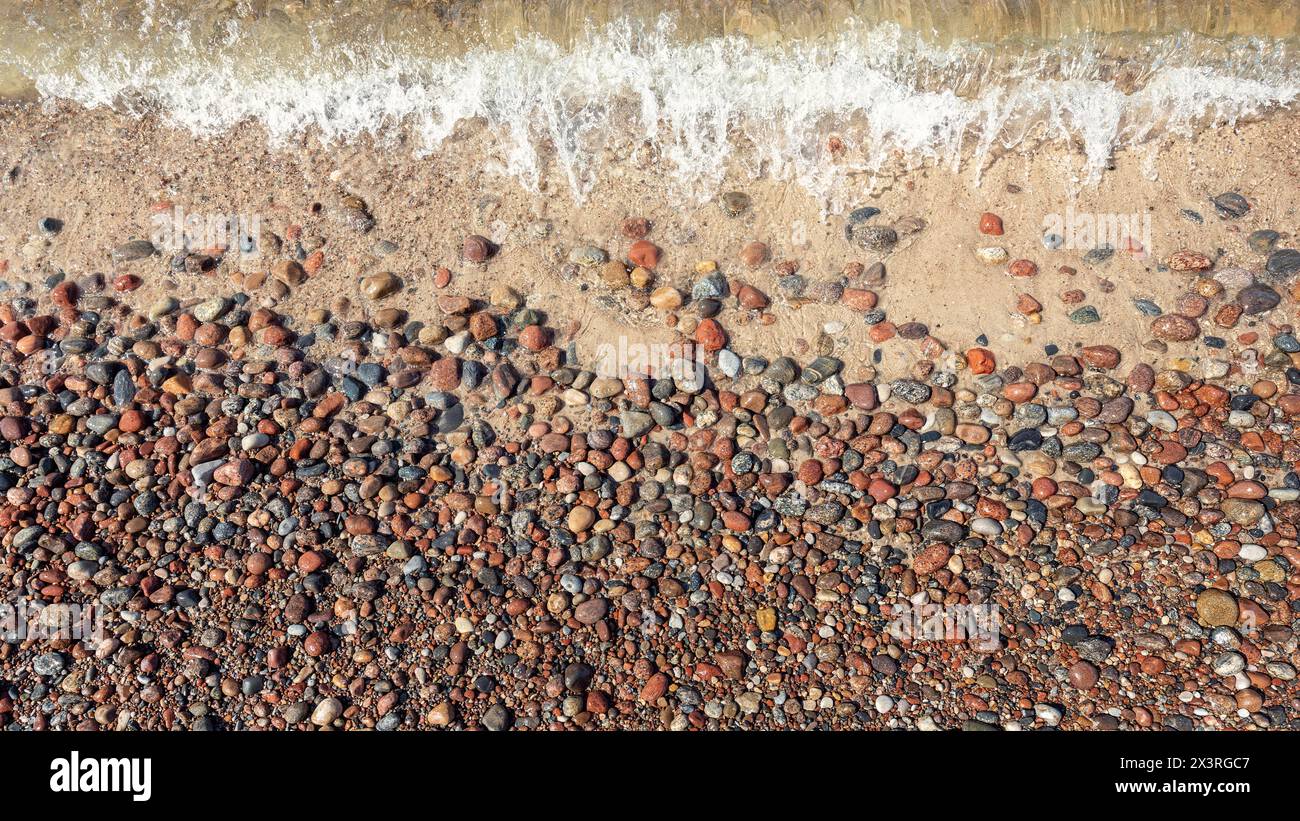 Vista piatta della riva con onde di mare spumeggianti e ciottoli rossi marroni lucidi. Foto Stock