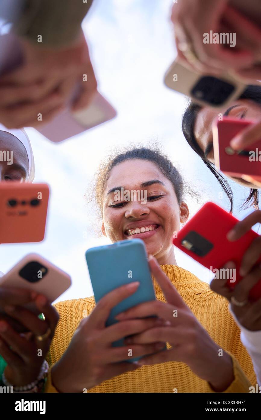 Angolo basso. Gruppo di giovani donne multirazziali sorridenti si riunirono in cerchio usando i telefoni cellulari Foto Stock