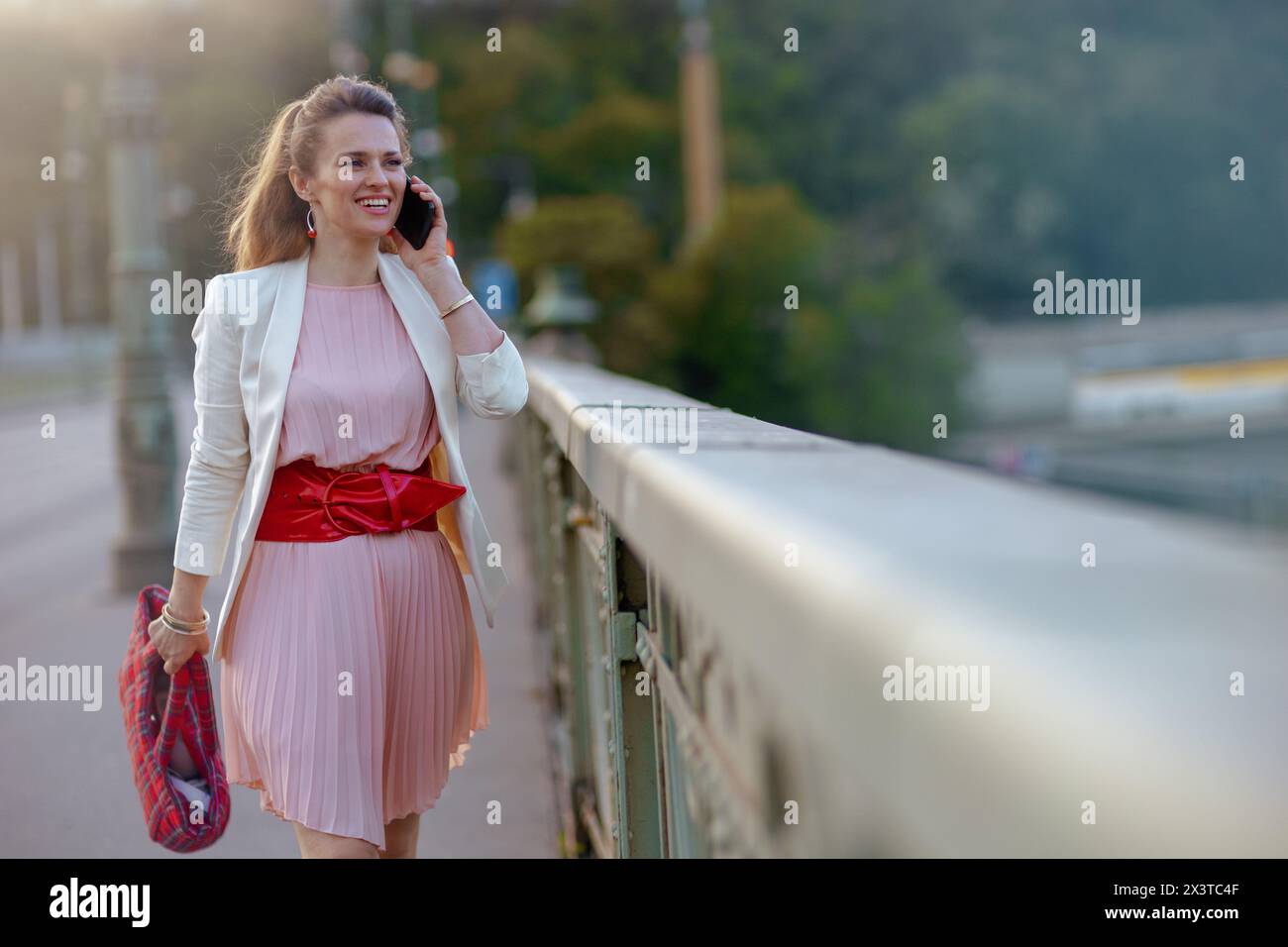 donna sorridente di 40 anni in abito rosa e giacca bianca in città utilizzando uno smartphone sul ponte. Foto Stock
