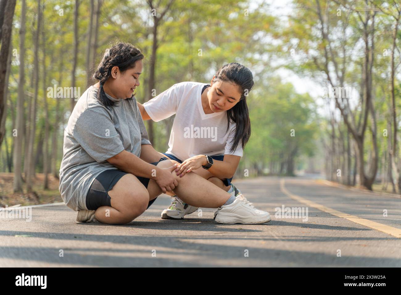 Donna in sovrappeso con lesioni al ginocchio seduta sulla pista da corsa in un parco di corsa tenendo il ginocchio doloroso mentre la sua amica di corsa seduta besi Foto Stock