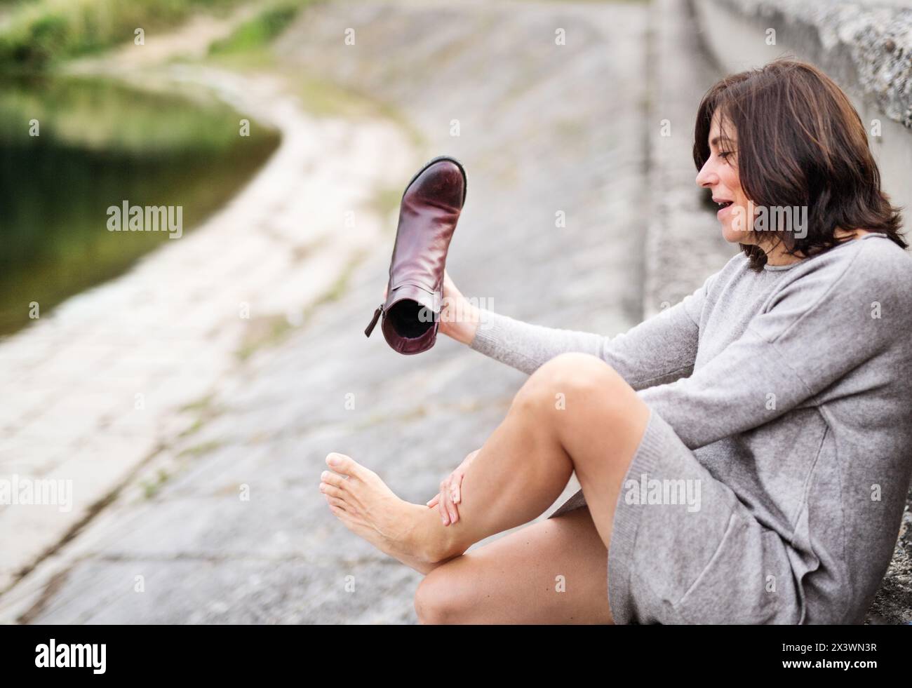 Donna che soffre di dolore dopo aver indossato stivali con tacco alto durante la passeggiata. Donna matura che massaggia le gambe doloranti, si toglie le scarpe strette. Foto Stock