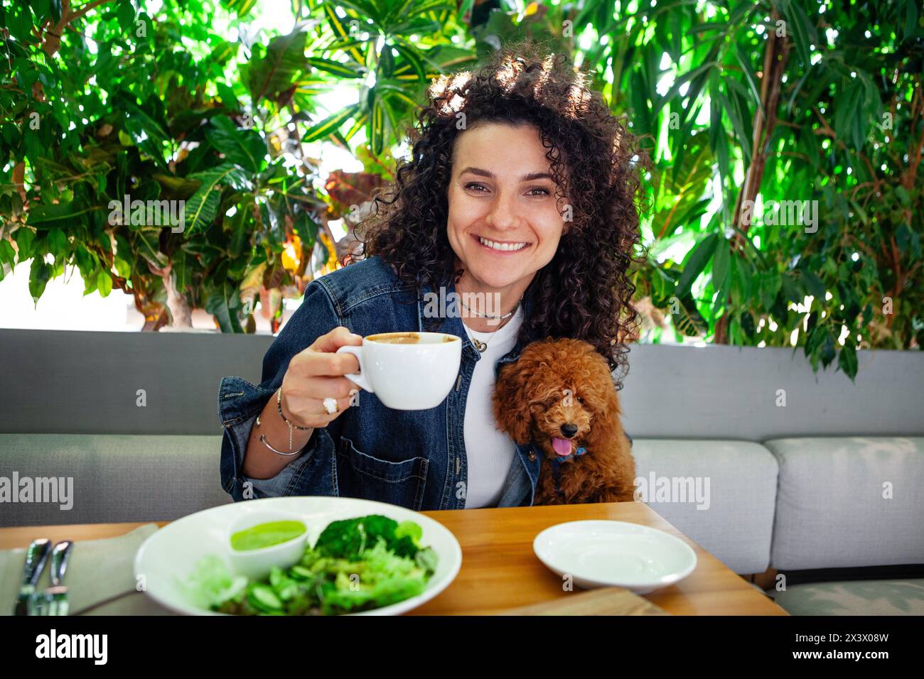 Ritratto di una bella donna bruna riccia con animale domestico che gusta un pasto di verdure a base di piante in un ristorante vegano con interni ecologici adornati con Foto Stock