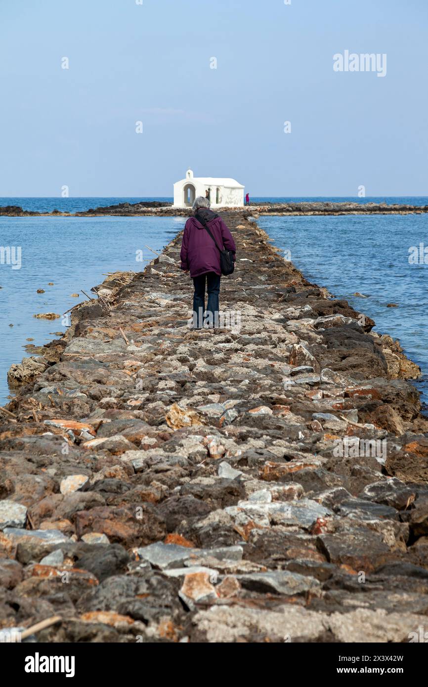 Un'anziana donna caucasica che cammina sul molo di pietra artificiale che conduce alla cappella bianca costruita sul mare di San Nicola (Agios Nikolaos) Foto Stock