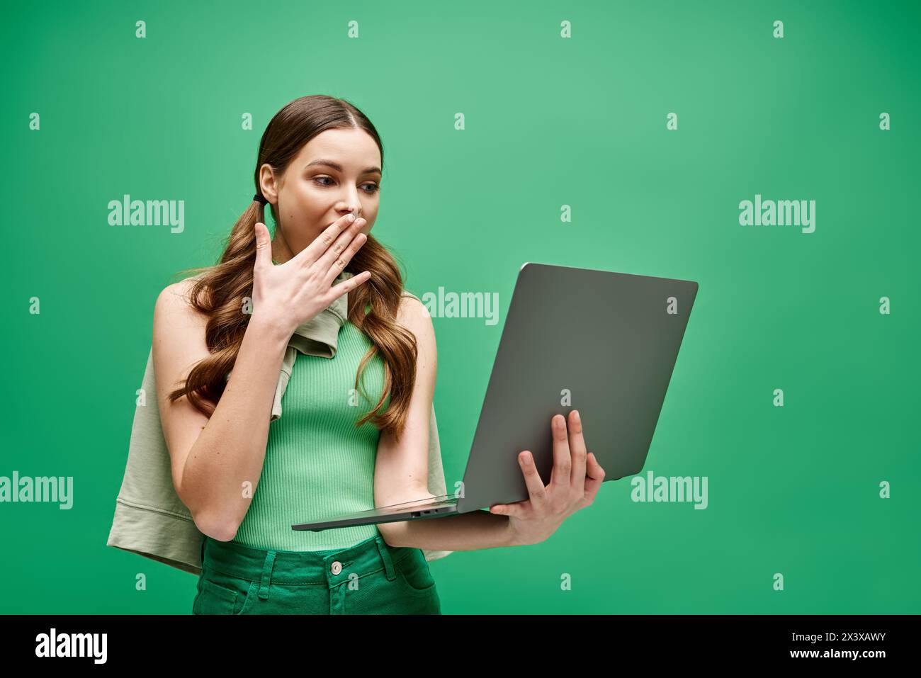 Una giovane donna di 20 anni si copre la bocca mentre usa un notebook in uno studio, suggerendo pensieri o emozioni nascosti. Foto Stock