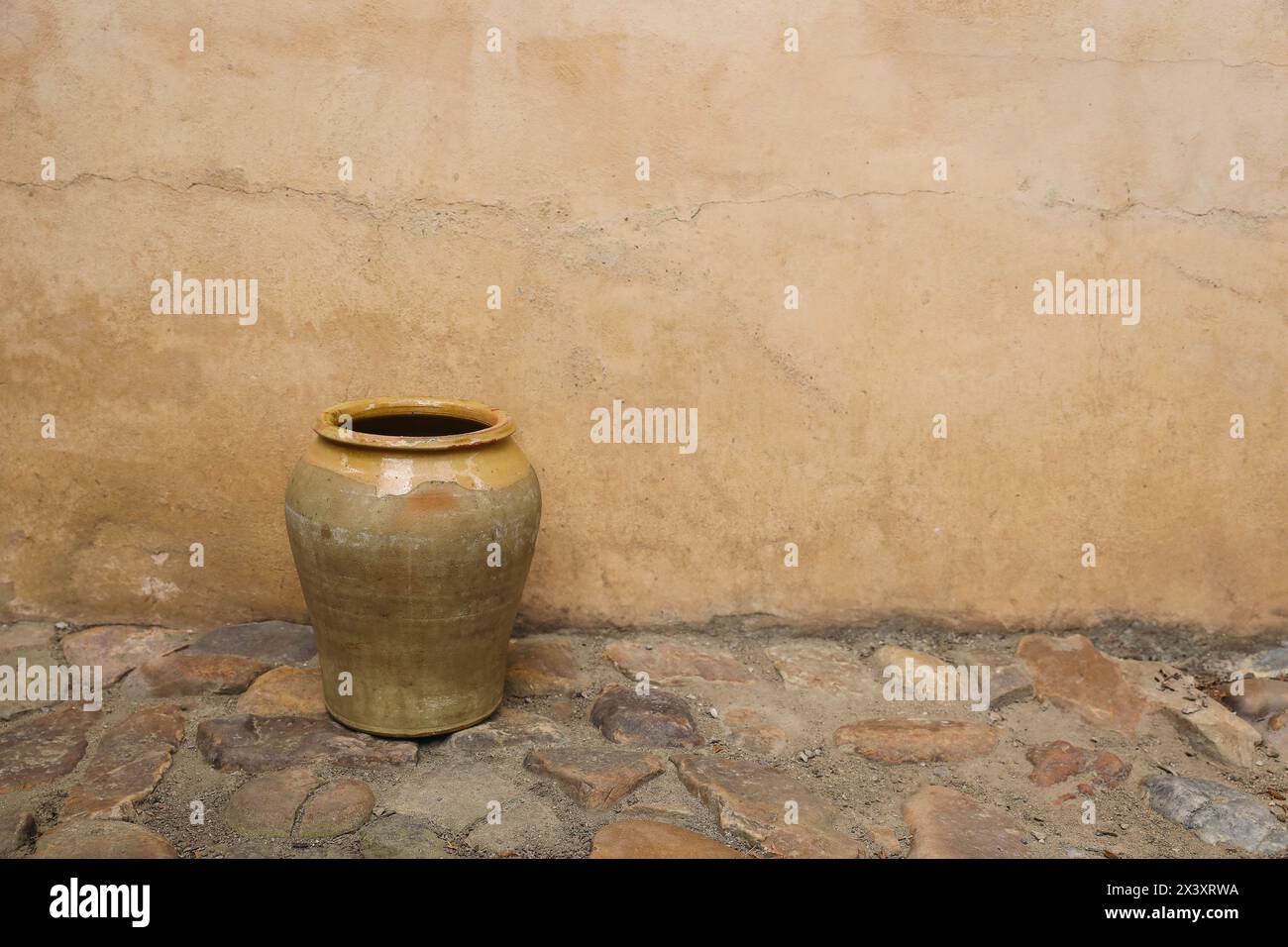 Vaso rustico in argilla gialla su pavimento in pietra di ciottoli, marciapiede contro muri scuri in ocra, facciata della casa. Vecchio vaso confit francese o spagnolo vintage Foto Stock