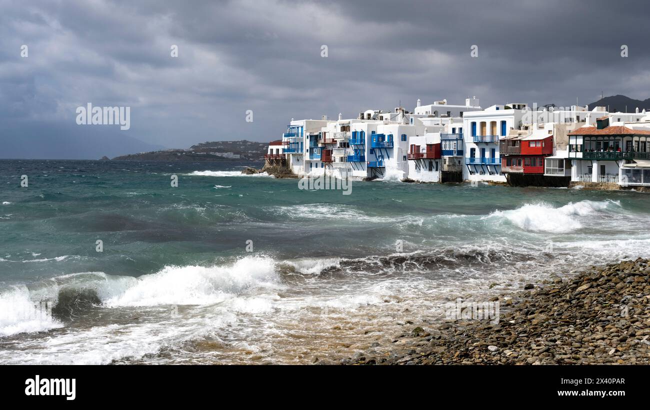 Edifici bianchi lungo la riva con un cielo tempestoso e onde che si infrangono sul lungomare dell'isola dell'Egeo meridionale di Mykonos; Mykonos, Grecia Foto Stock