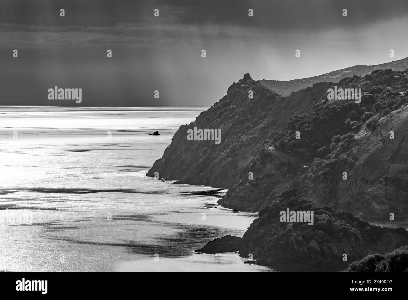 Immagine in bianco e nero di una vista costiera delle scogliere sarde che si affacciano sul Mar Mediterraneo con i raggi del sole che brillano attraverso le nuvole scure Foto Stock