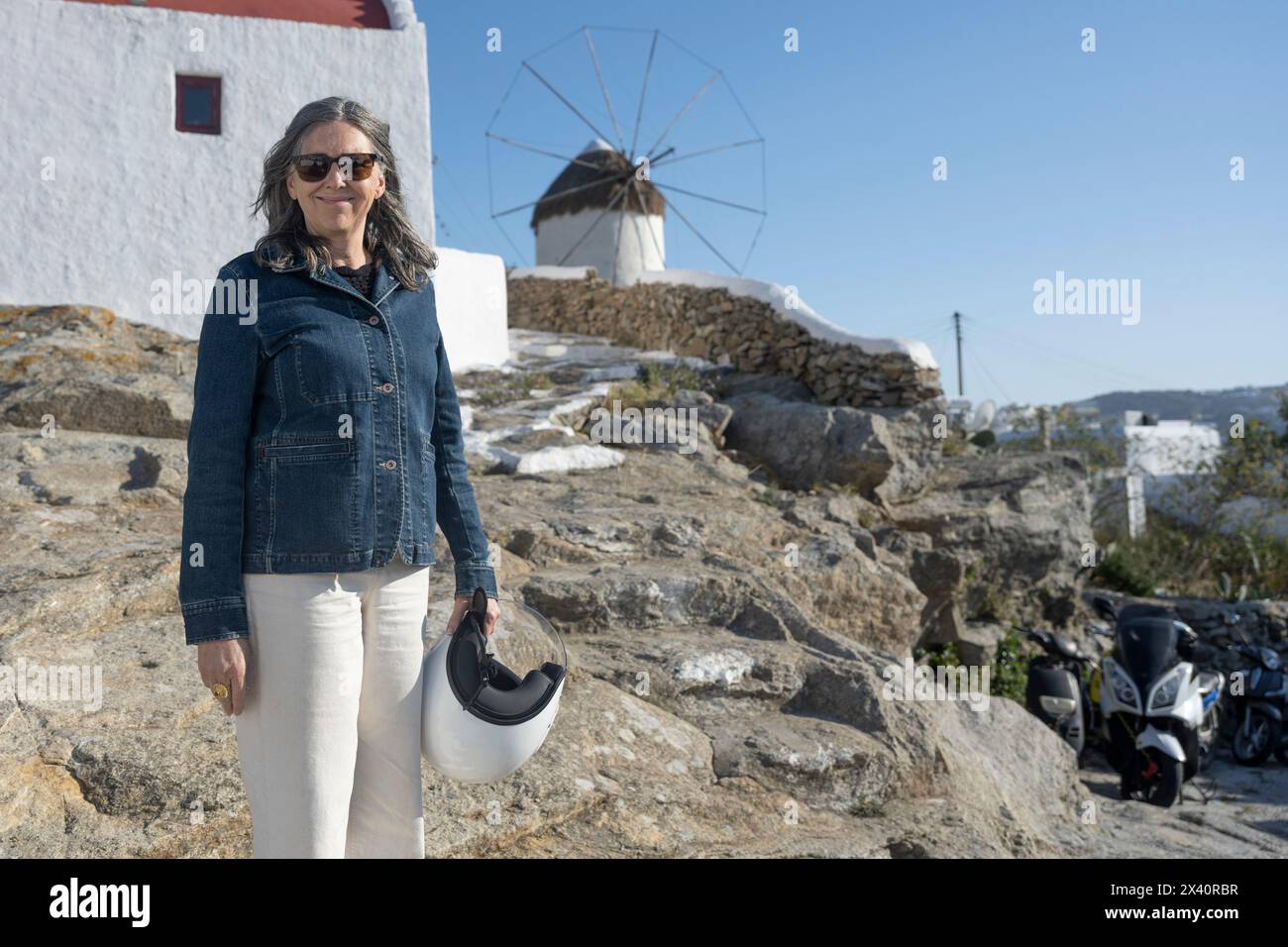 Donna anziana con casco posa in un mulino a vento a Mykonos, Grecia; Mykonos, Egeo meridionale, Grecia Foto Stock