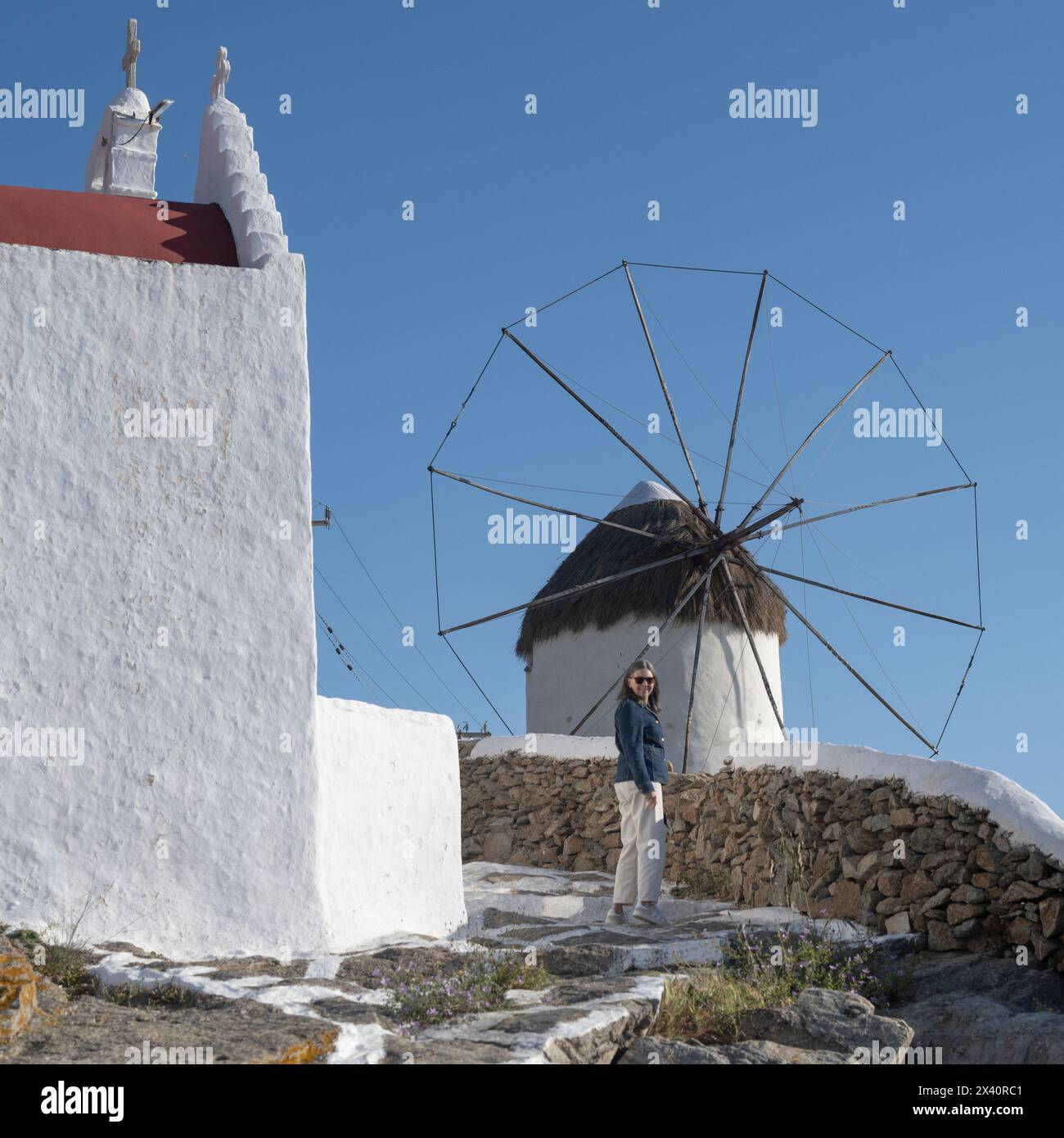 Donna anziana in piedi fuori da una chiesa e un mulino a vento a Mykonos, Grecia; Mykonos, Egeo meridionale, Grecia Foto Stock