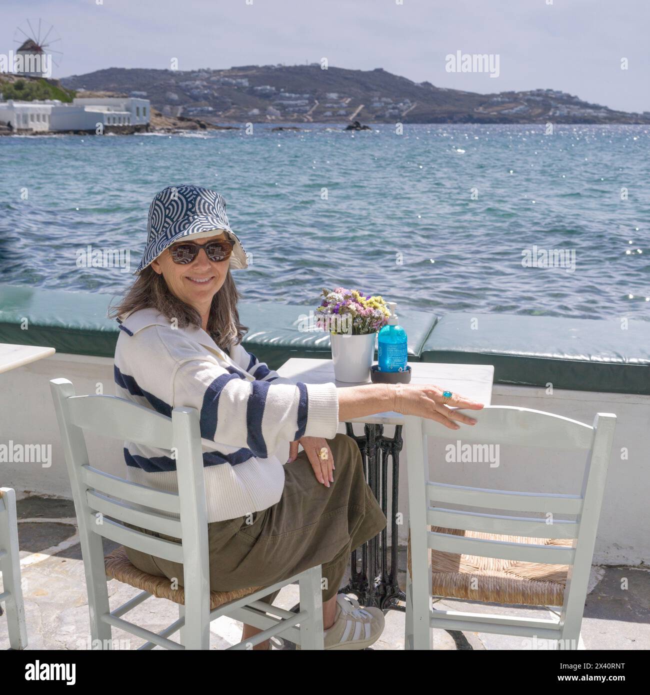 Una donna che indossa un cappello da sole e un paio di occhiali da sole gode di un caffè sul lungomare in una giornata di sole a Mykonos, Grecia; Mykonos, Grecia Foto Stock