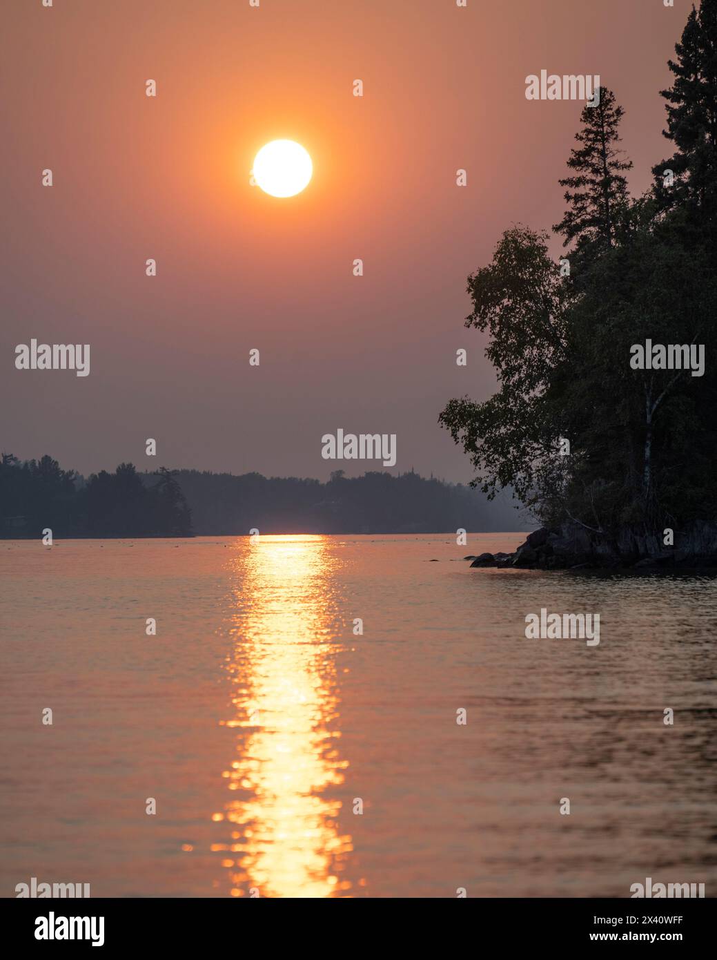 Una spettacolare luce del tramonto su un lago con alberi sagomati lungo la costa e un raggio di sole riflesso sulla superficie dell'acqua Foto Stock
