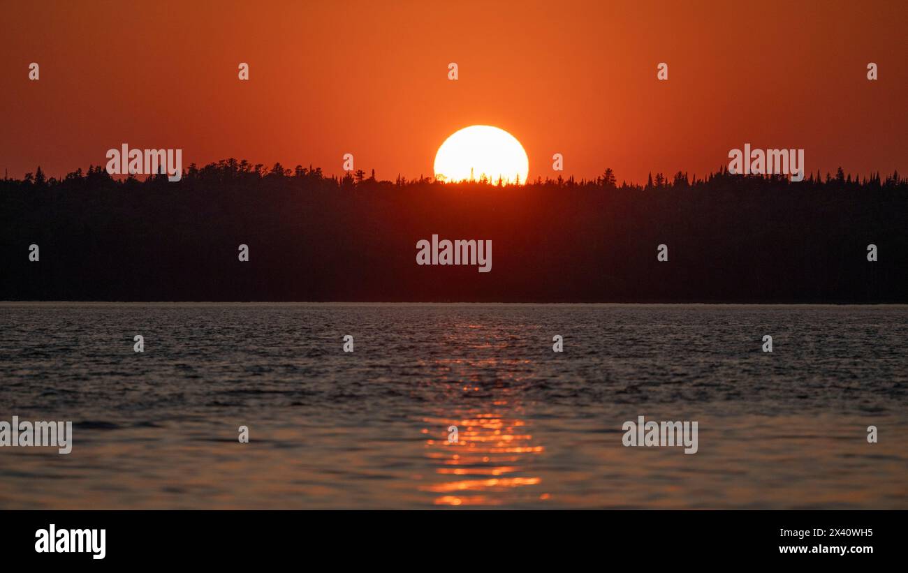 Sole incandescente che tramonta su un lago con alberi sagomati lungo la costa e un raggio di sole riflesso sulla superficie dell'acqua Foto Stock