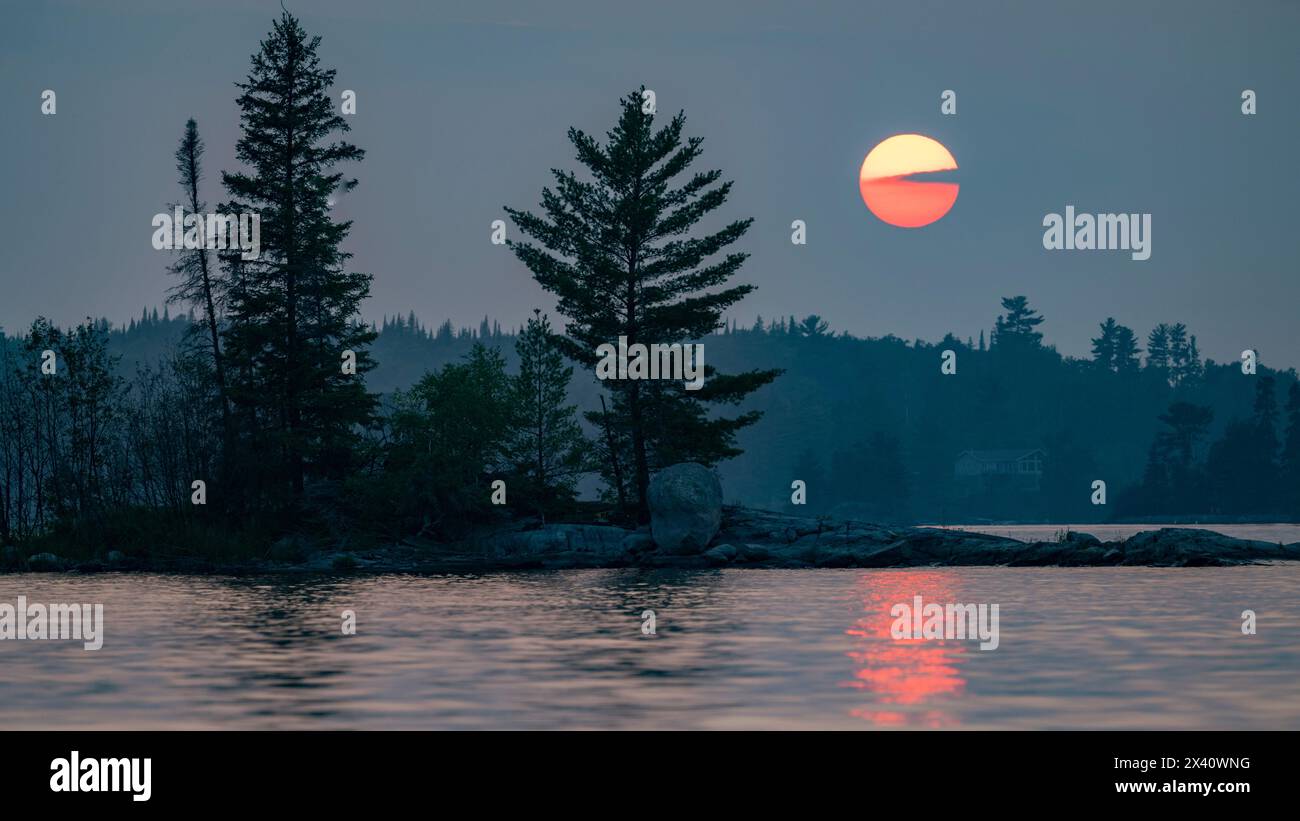 Sole incandescente che tramonta su un lago con alberi sagomati lungo la costa e un raggio di sole riflesso sulla superficie dell'acqua Foto Stock