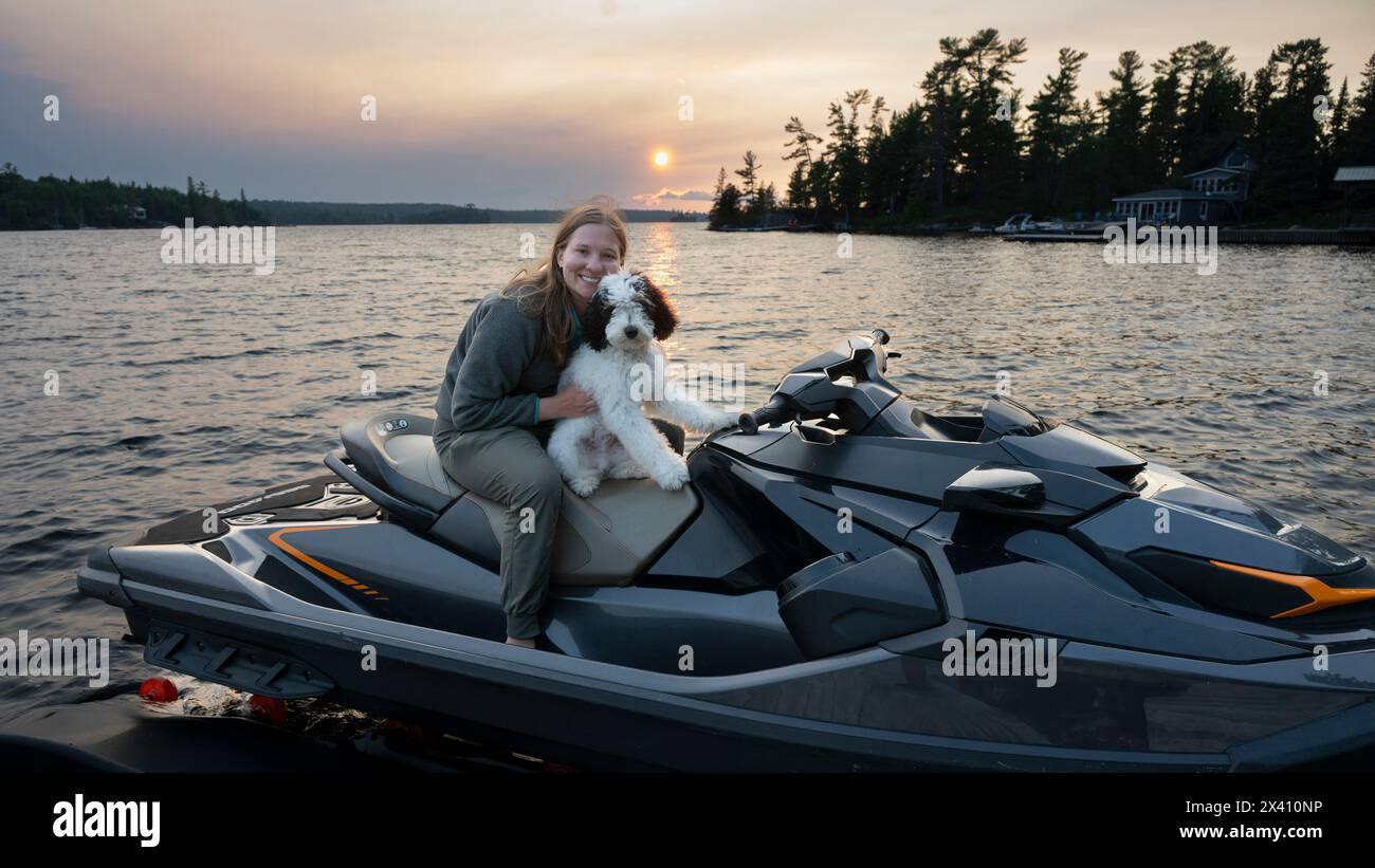 Una giovane donna tiene il suo cane su una moto d'acqua personale su un lago al tramonto, in posa per la macchina fotografica; Lake of the Woods, Ontario, Canada Foto Stock