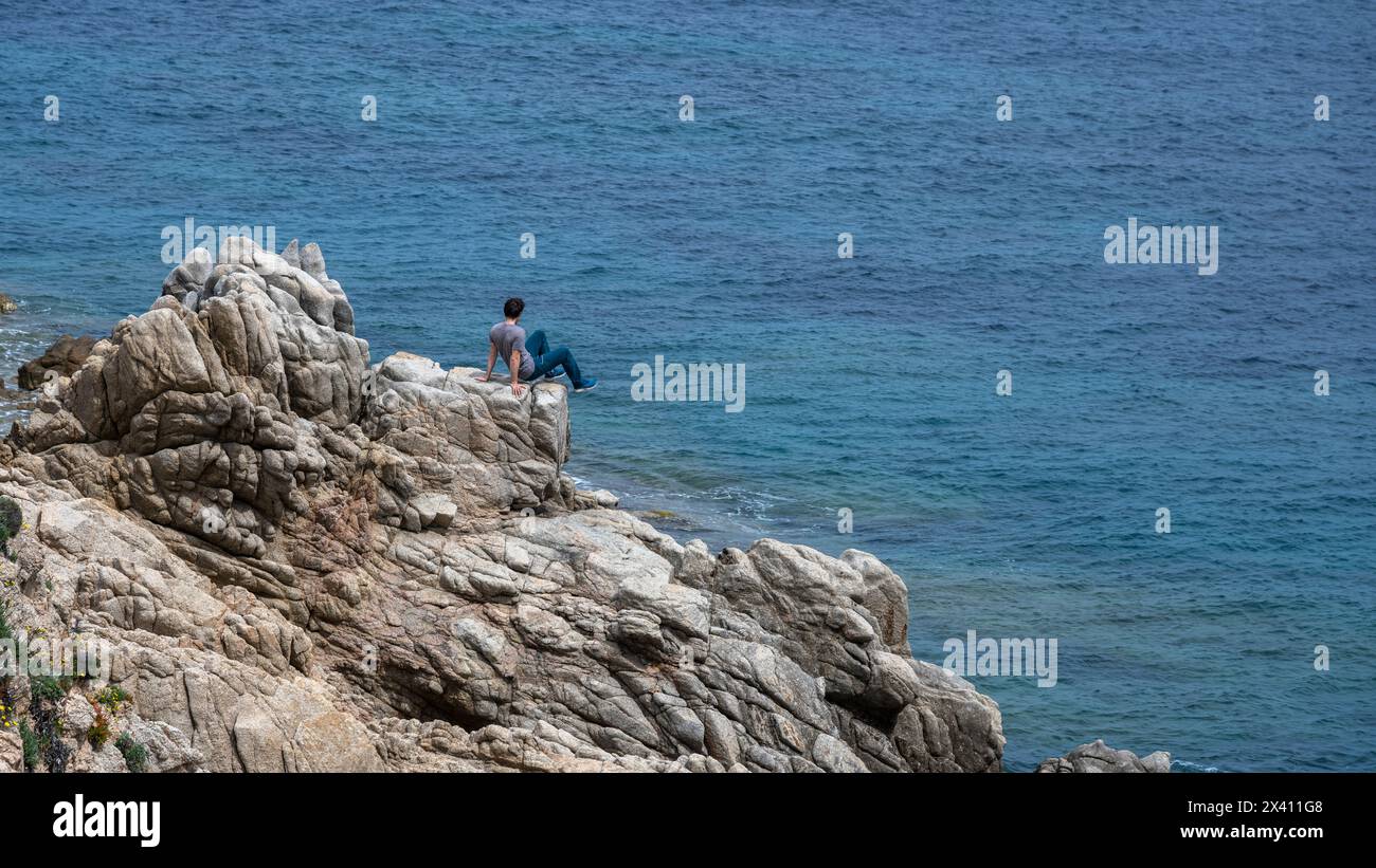 L'uomo siede su una costa rocciosa frastagliata con vista sul blu del Mar Mediterraneo; Santa Teresa Gallura, Sassari, Italia Foto Stock