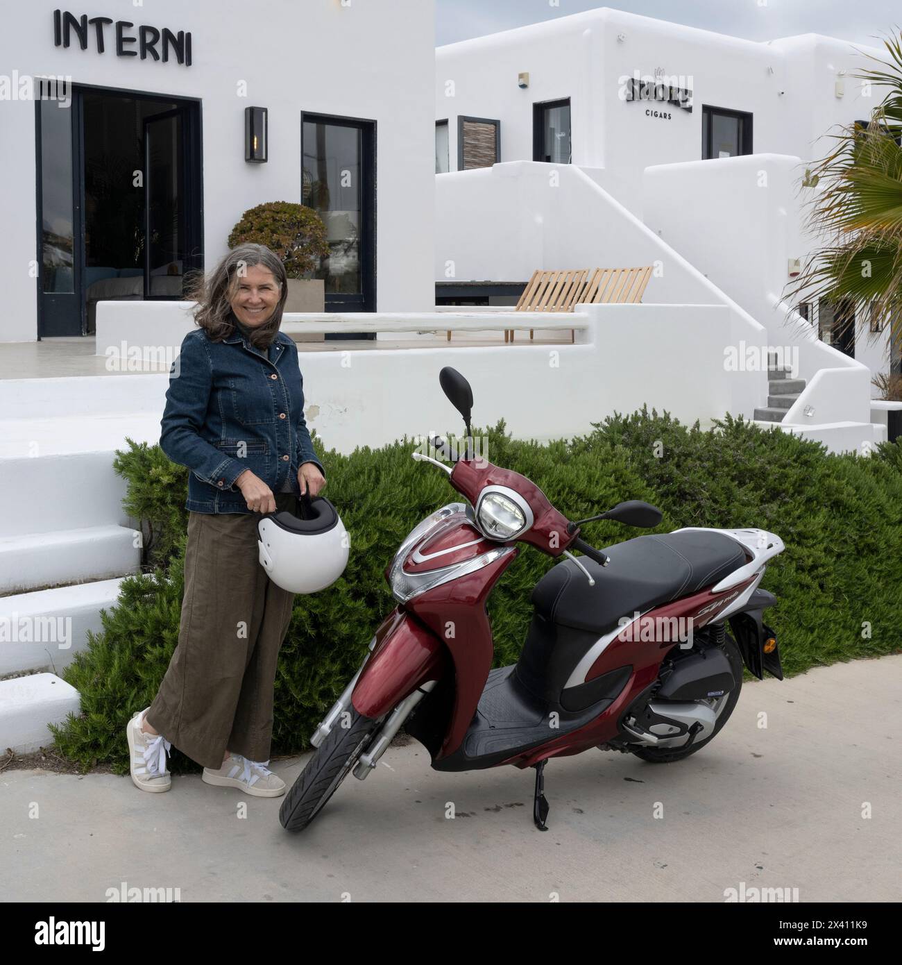 Donna sta con la sua moto all'esterno degli edifici bianchi di Mykonos, Grecia; Mykonos, Egeo meridionale, Grecia Foto Stock