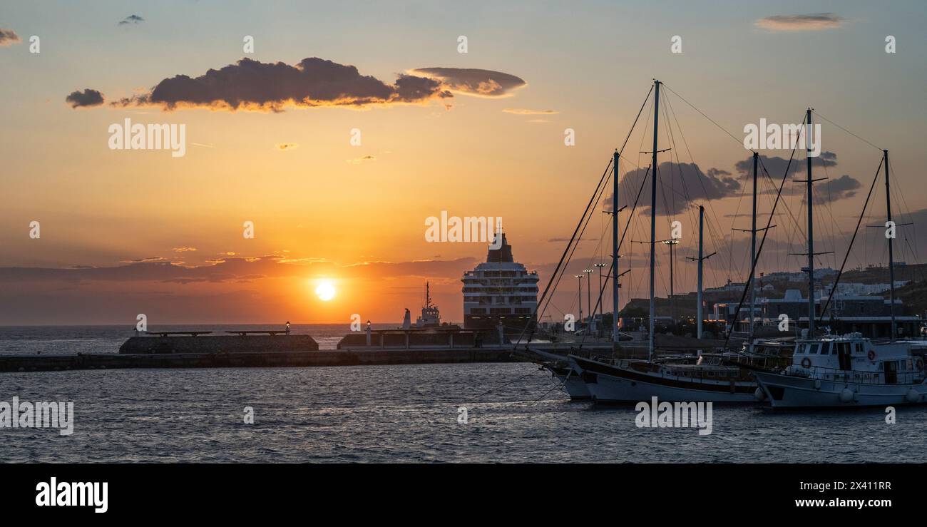 Nave da crociera nel porto di Mykonos con un sole luminoso al tramonto e barche a vela nel porto in primo piano; Mykonos, Egeo meridionale, Grecia Foto Stock