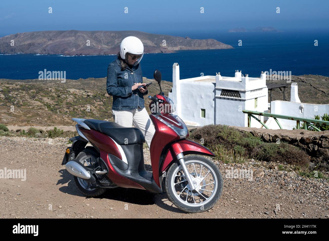 Una donna in viaggio in moto controlla il suo smartphone con una vista sulla costa e sul Mar Egeo; Ano Mera, Egeo meridionale, Grecia Foto Stock