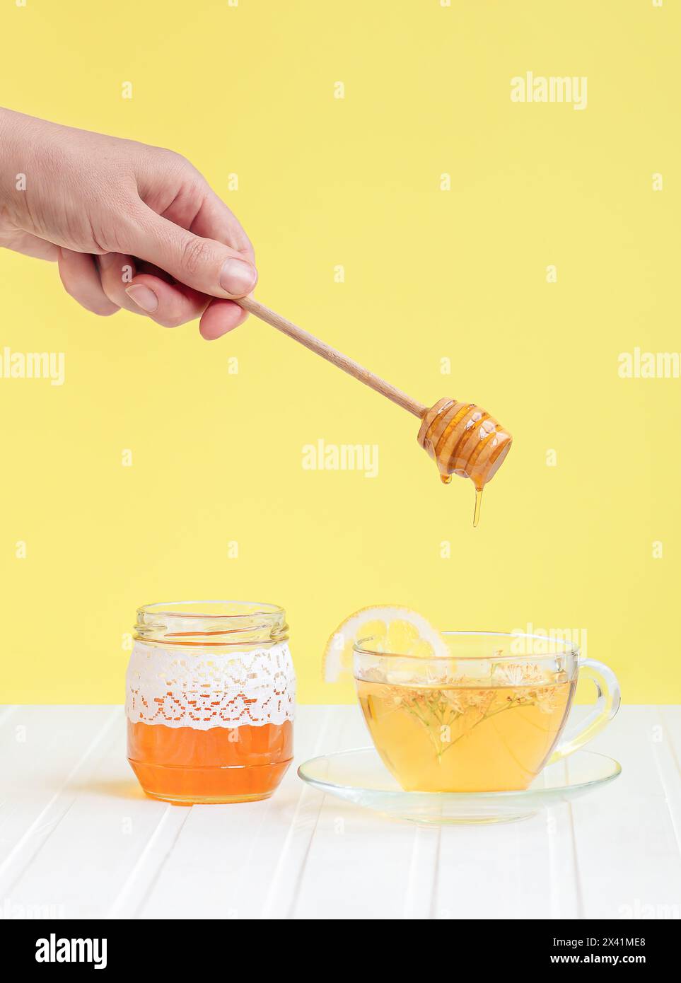 Fai gocciolare il miele su una tazza di tè alle erbe circondato da fiori e bottiglie. Concetto di stile di vita sano e rimedi naturali Foto Stock