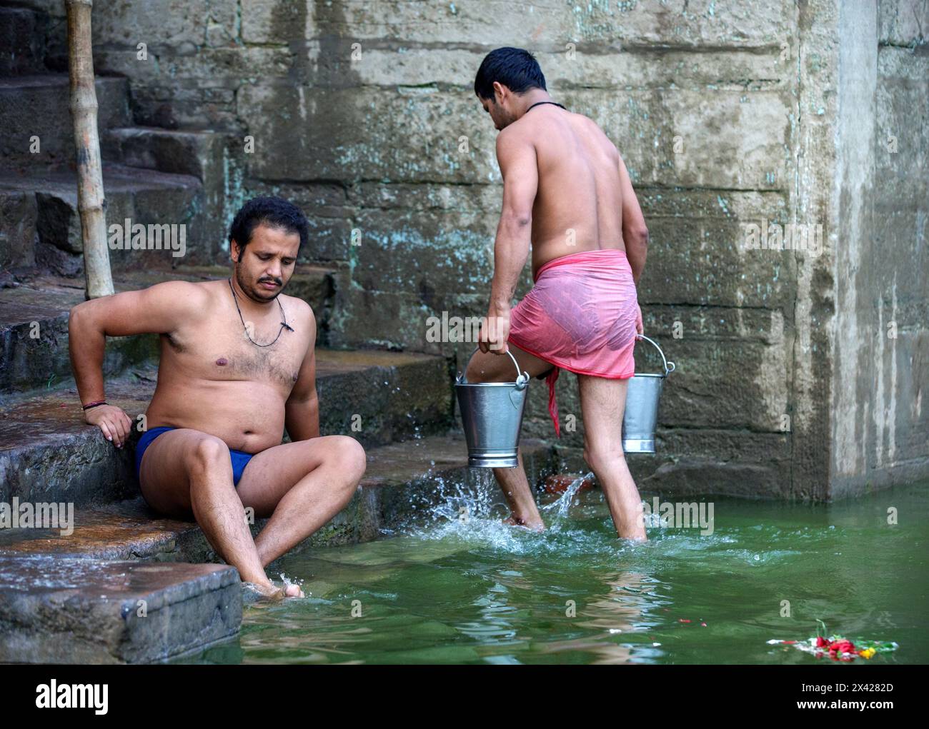 Due uomini con i piedi nel fiume Gange a Varanasi, in India, uno con due secchi di metallo di acqua del Gange. Foto Stock