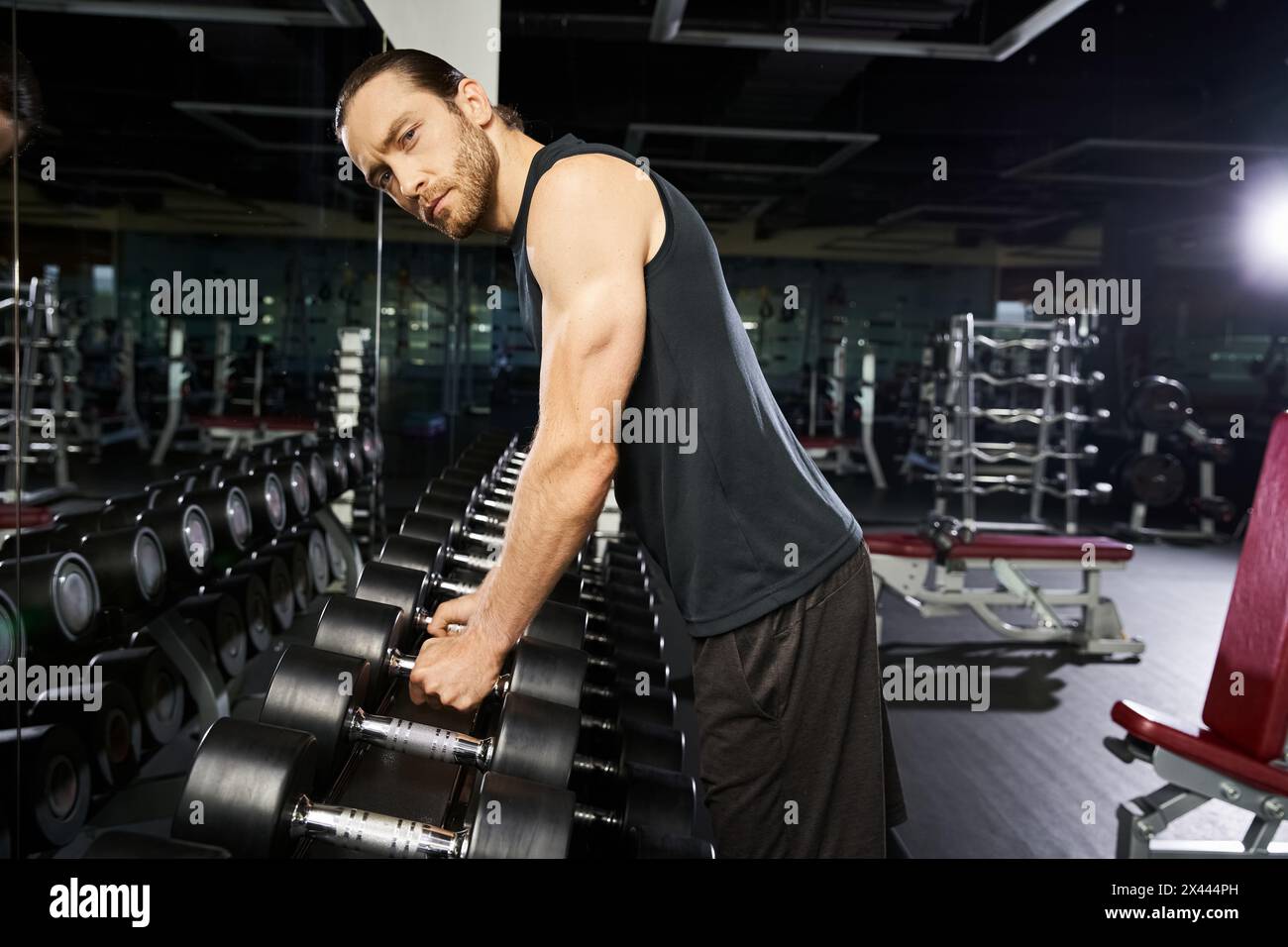 Un uomo aderente all'abbigliamento sportivo si trova accanto a una fila di manubri in palestra, preparandosi per un'intensa sessione di allenamento. Foto Stock