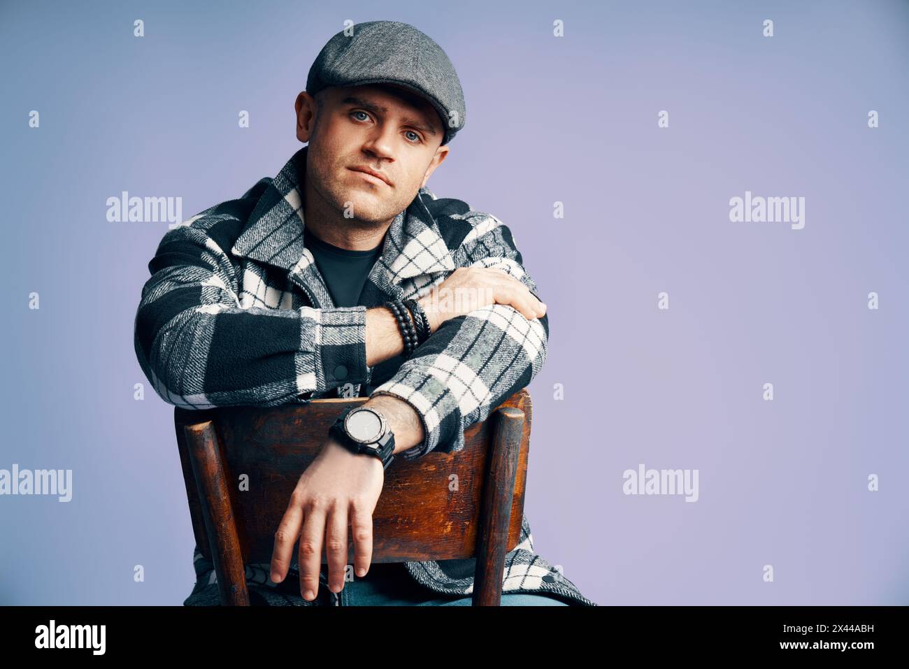 Uomo elegante e affascinante che posa seduto sulla sedia sullo sfondo blu dello studio. Ispirazione in stile Peaky Blinders Foto Stock