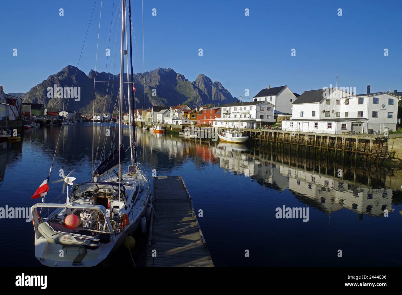 Le navi a vela e le case di legno si riflettono nelle acque calme di un piccolo porto, Rorbuer, Idyll, Henningsvaer, Vagan, Nordland, Norvegia Foto Stock
