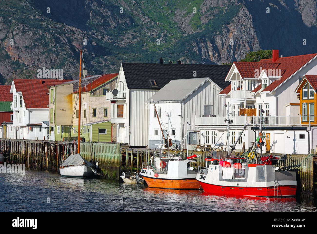 Barche da pesca di fronte a case di legno e alte montagne in un piccolo porto, Idyll, Henningsvaer, Vagan, Nordland, Norvegia Foto Stock