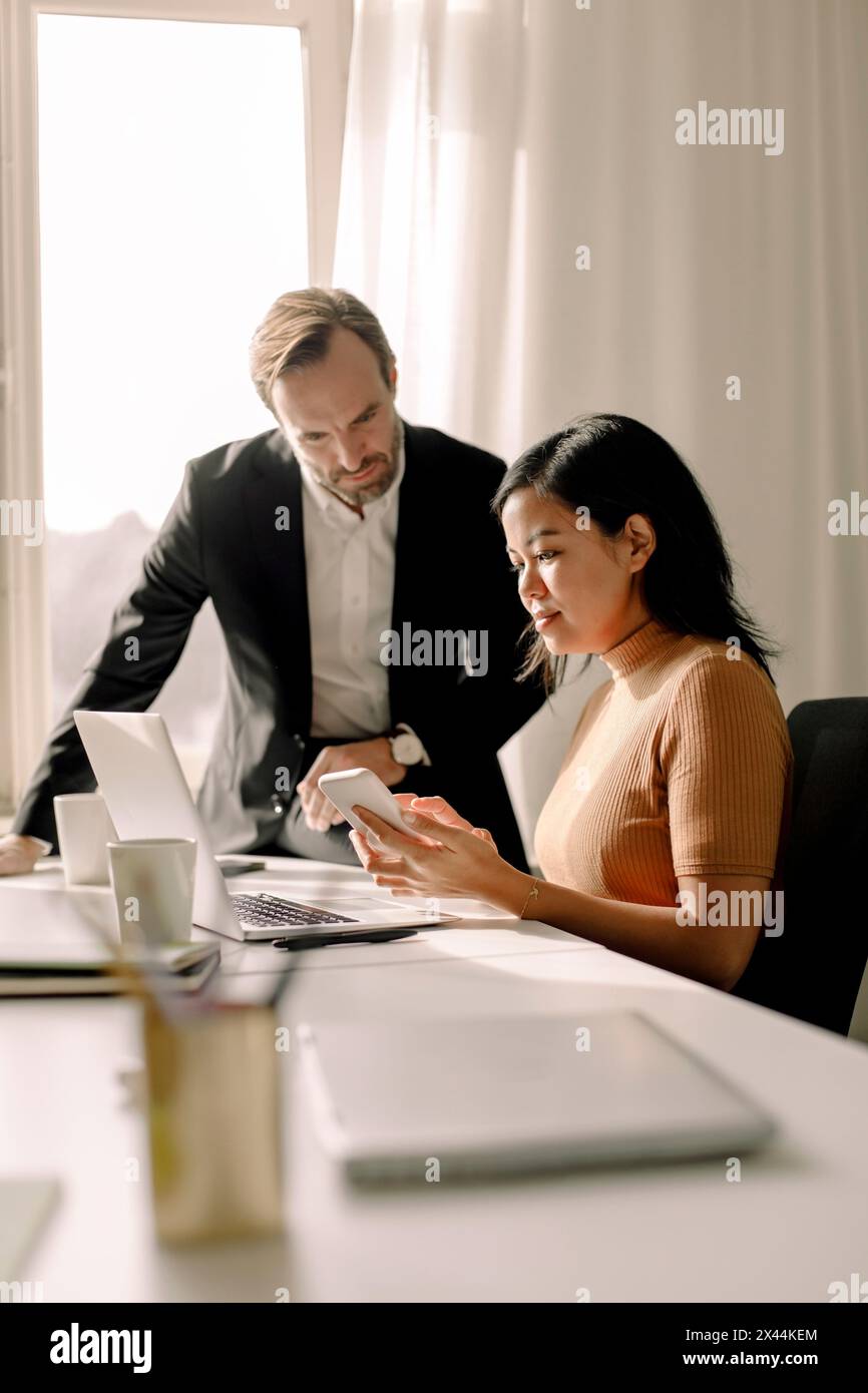 Donna d'affari che condivide lo smartphone con un uomo d'affari in ufficio Foto Stock