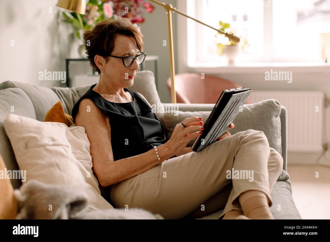 Donna anziana in pensione che utilizza un tablet digitale mentre si siede sul divano di casa Foto Stock