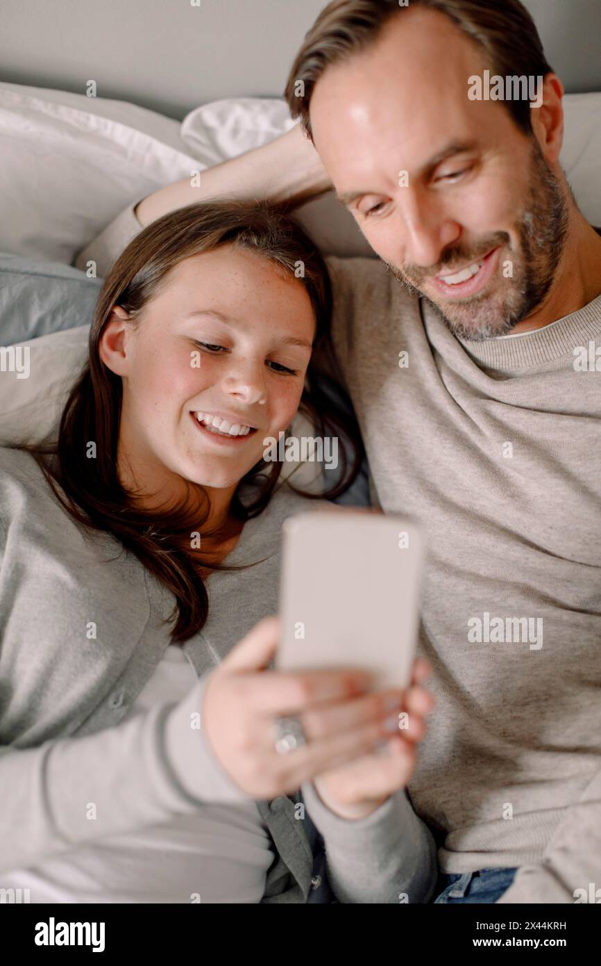 Felice ragazza adolescente che condivide lo smartphone con il padre mentre è sdraiata a letto Foto Stock