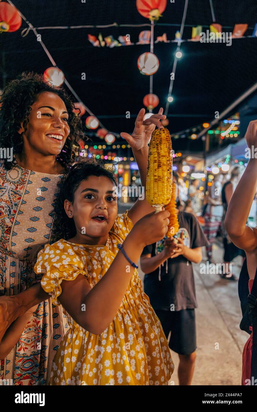 Ragazza eccitata che tiene il mais alla griglia mentre si trova accanto alla madre al mercato alimentare in vacanza Foto Stock