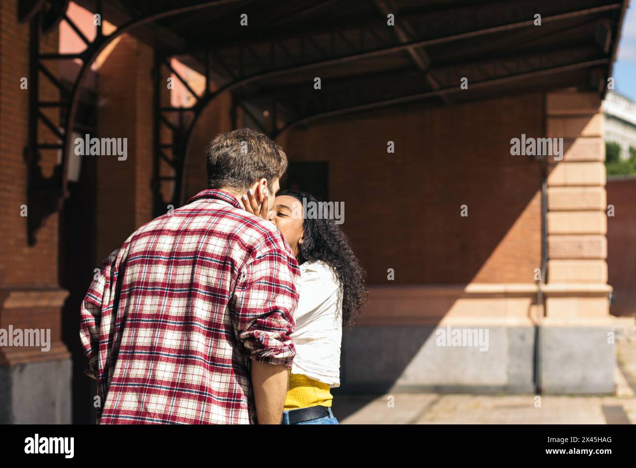 Giovane coppia multirazziale che abbraccia e bacia dopo l'incontro di nuovo alla stazione. Sta portando con sé una valigia blu dal suo viaggio e lui aspetta Foto Stock