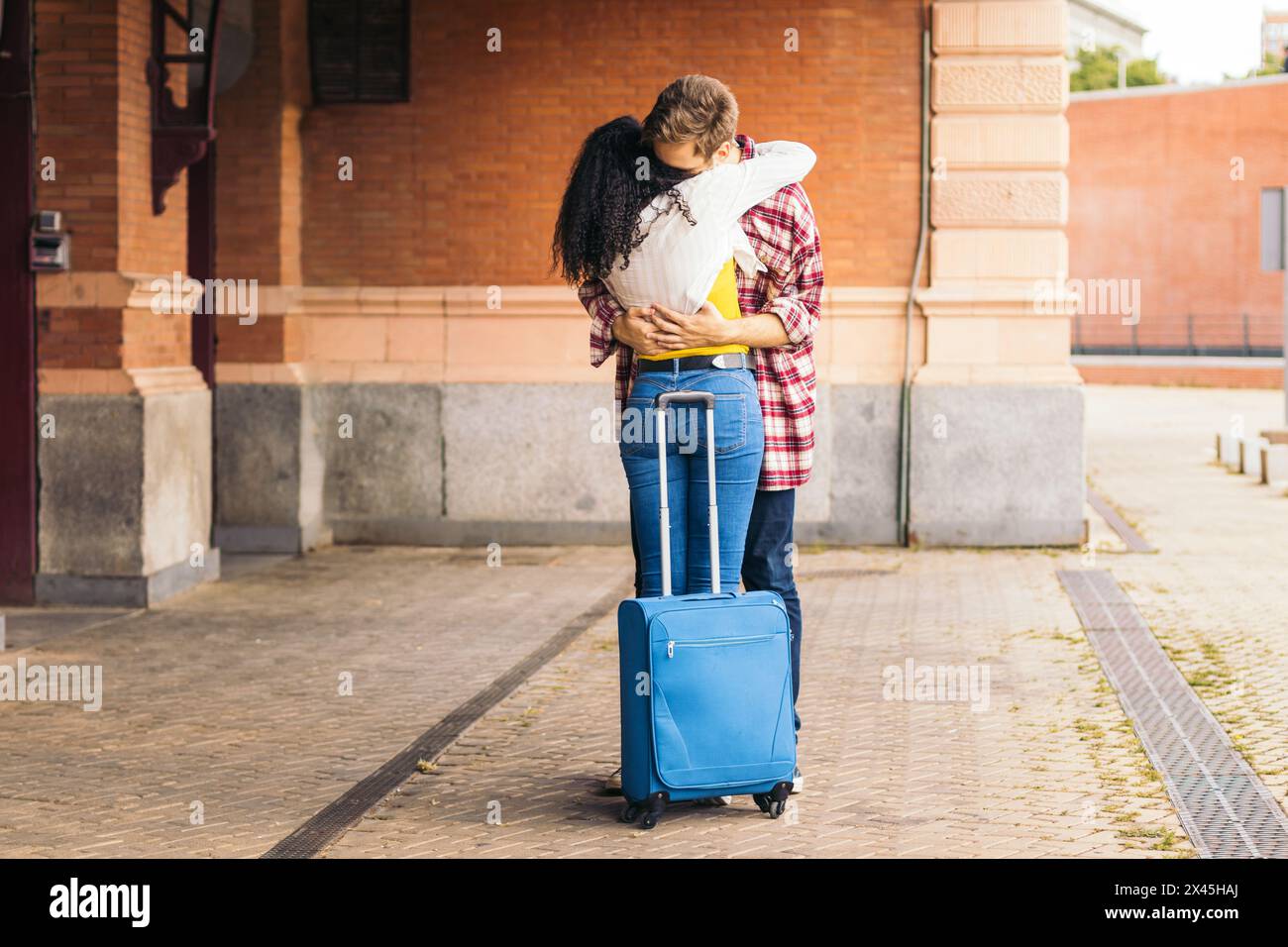 Giovane coppia multirazziale che abbraccia e bacia dopo l'incontro di nuovo alla stazione. Sta portando con sé una valigia blu dal suo viaggio e lui aspetta Foto Stock