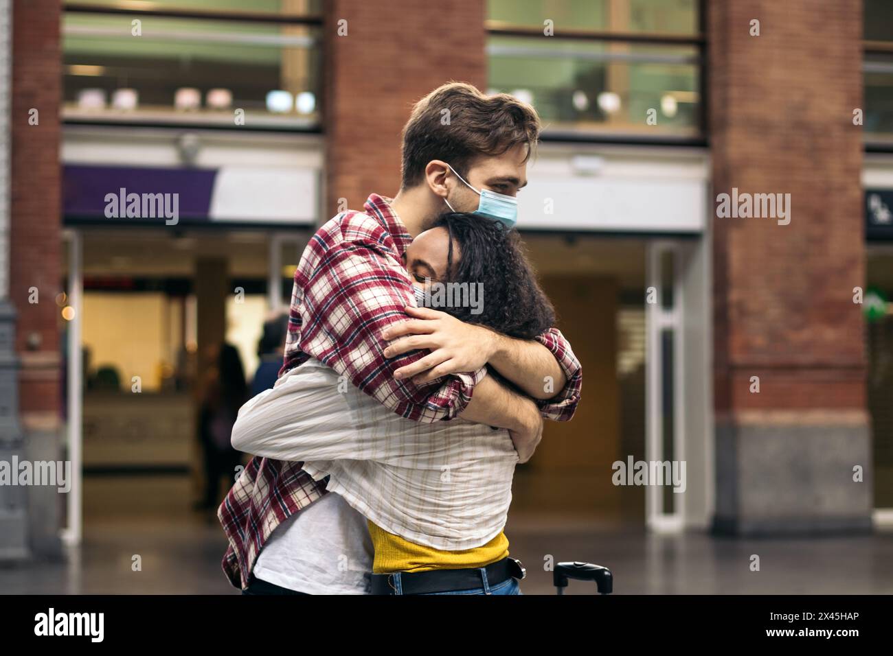 Giovane coppia multirazziale che abbraccia e bacia dopo l'incontro di nuovo alla stazione. Sta portando con sé una valigia blu dal suo viaggio e lui aspetta Foto Stock