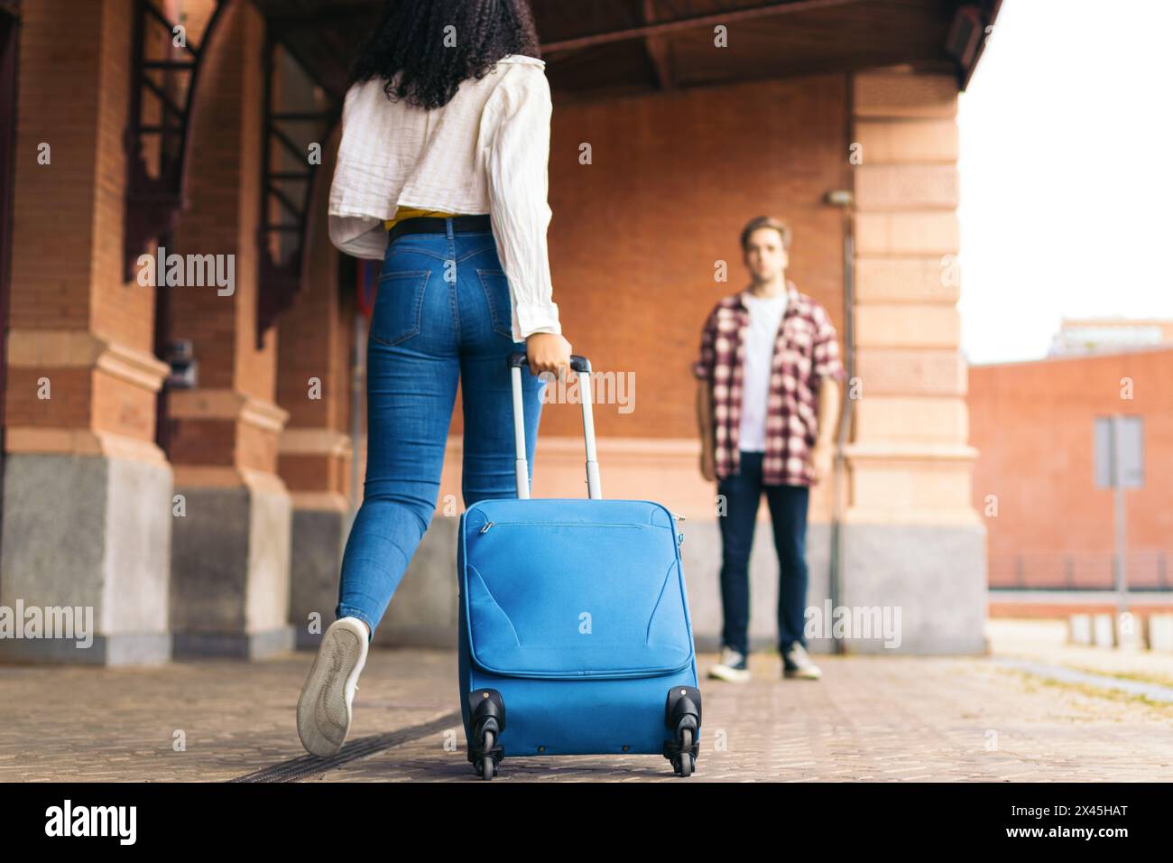 Giovane coppia multirazziale che abbraccia e bacia dopo l'incontro di nuovo alla stazione. Sta portando con sé una valigia blu dal suo viaggio e lui aspetta Foto Stock