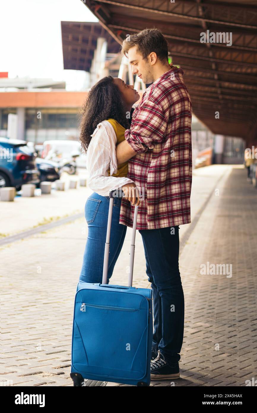 Giovane coppia multirazziale che abbraccia e bacia dopo l'incontro di nuovo alla stazione. Sta portando con sé una valigia blu dal suo viaggio e lui aspetta Foto Stock