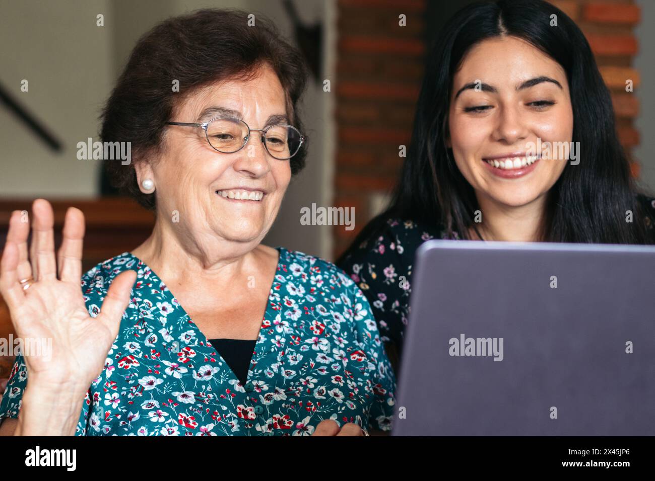 Una donna anziana sorridente che utilizza un laptop a casa e fa una videochiamata insieme a sua nipote. La nonna ondeggia alla telecamera Foto Stock