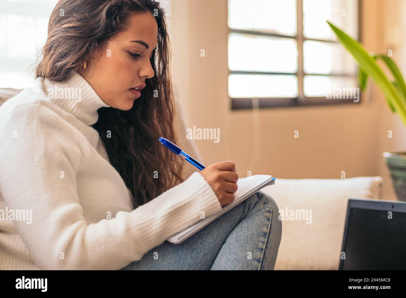 Giovane donna seduta a casa sua, che studia per l'università con il suo portatile e un notebook. Lo studente prende appunti durante la revisione delle informazioni Foto Stock